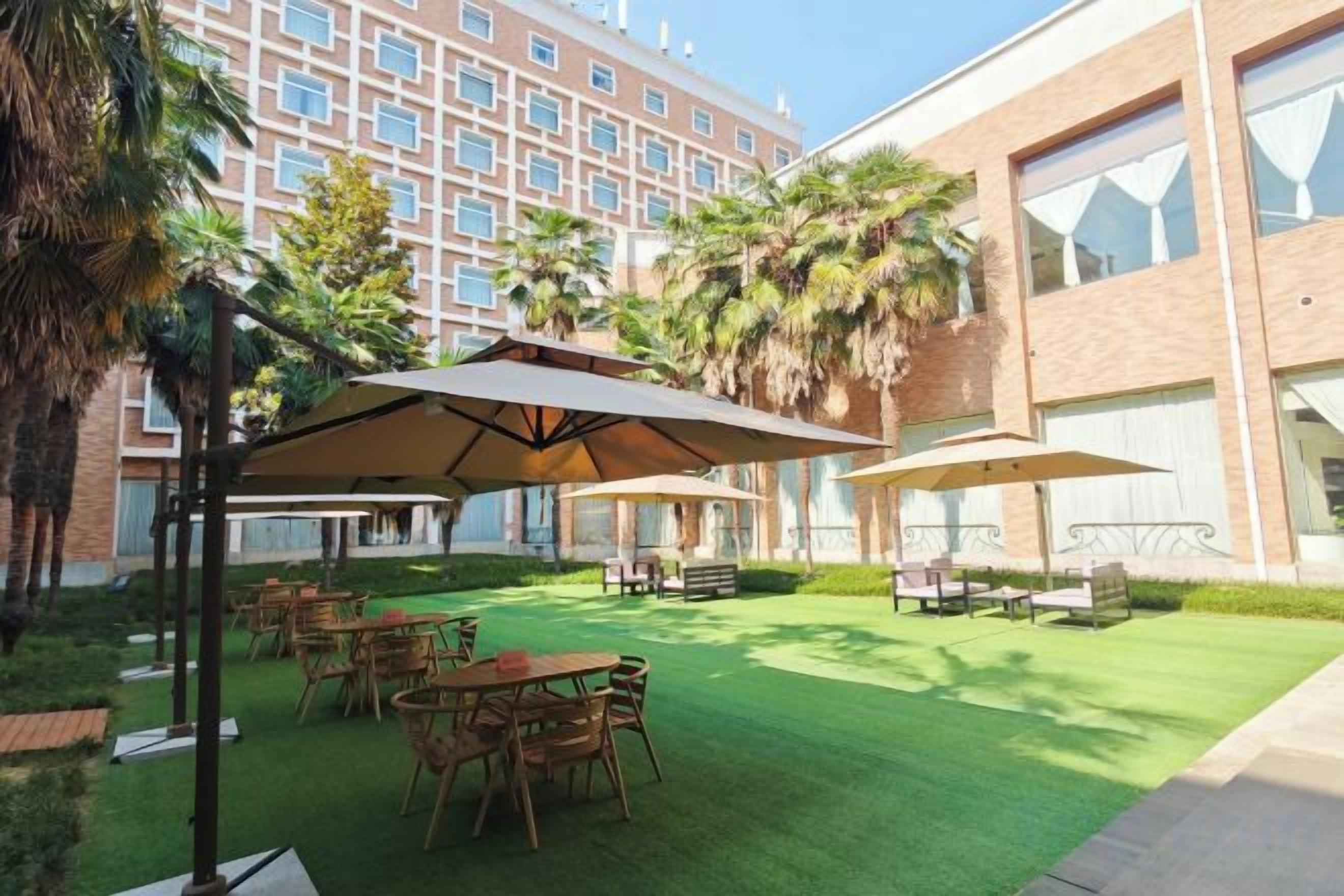 An outdoor patio area with tables and umbrellas, surrounded by greenery and a building in the background.
