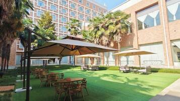 An outdoor patio area with tables and umbrellas, surrounded by greenery and a building in the background.