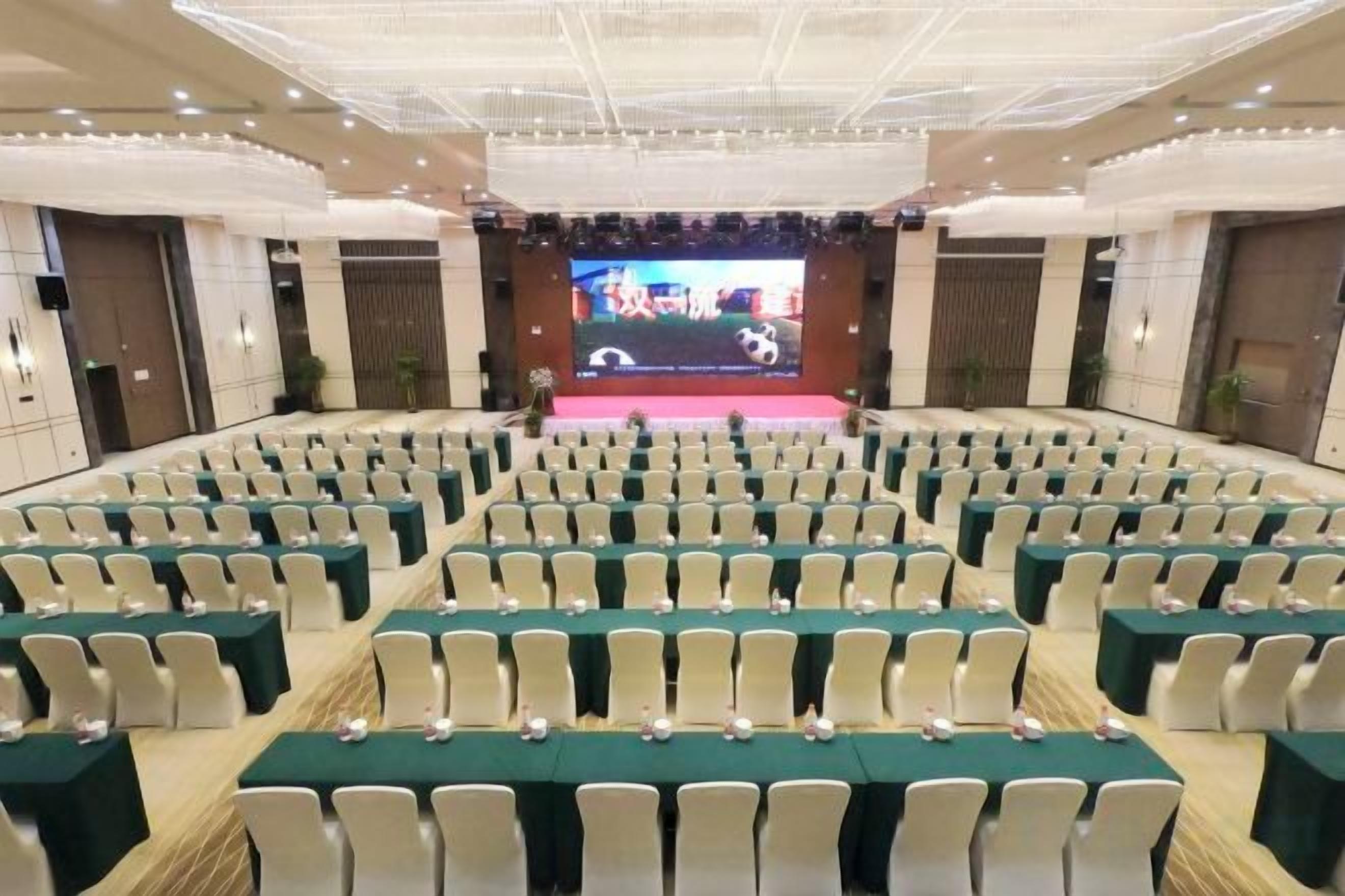 A conference room with rows of tables and chairs, a stage with a large screen, and a ceiling with recessed lighting.