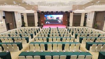 A conference room with rows of tables and chairs, a stage with a large screen, and a ceiling with recessed lighting.