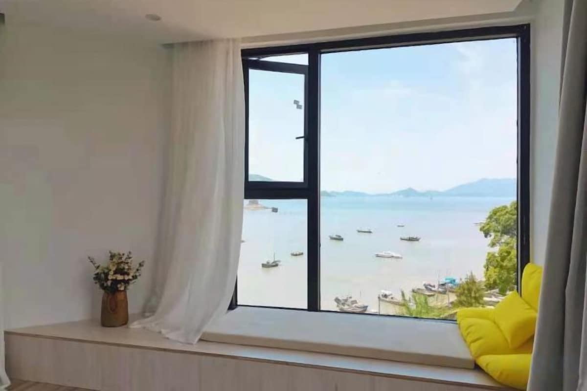 A room with a large window overlooking a body of water with boats, a yellow chair, and a small plant on a windowsill.