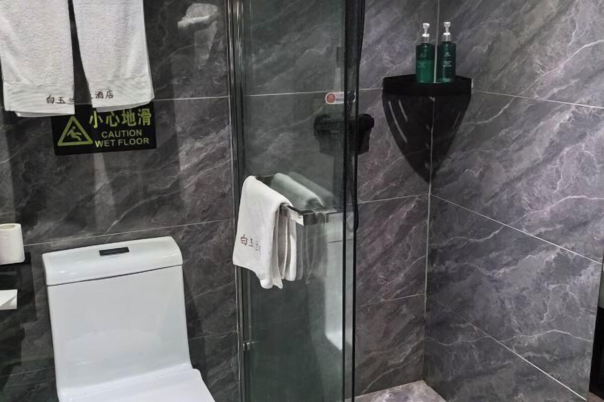 Casa de banho com chuveiro em cabine de vidro, sanita branca e prateleira preta com artigos de higiene pessoal.