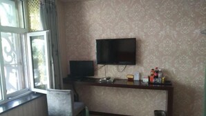 A room with a TV mounted on the wall, a desk with a computer and various items, a chair, and a window with a view outside. - Hailin Xinjia Fashion Express Hotel (Hailin)