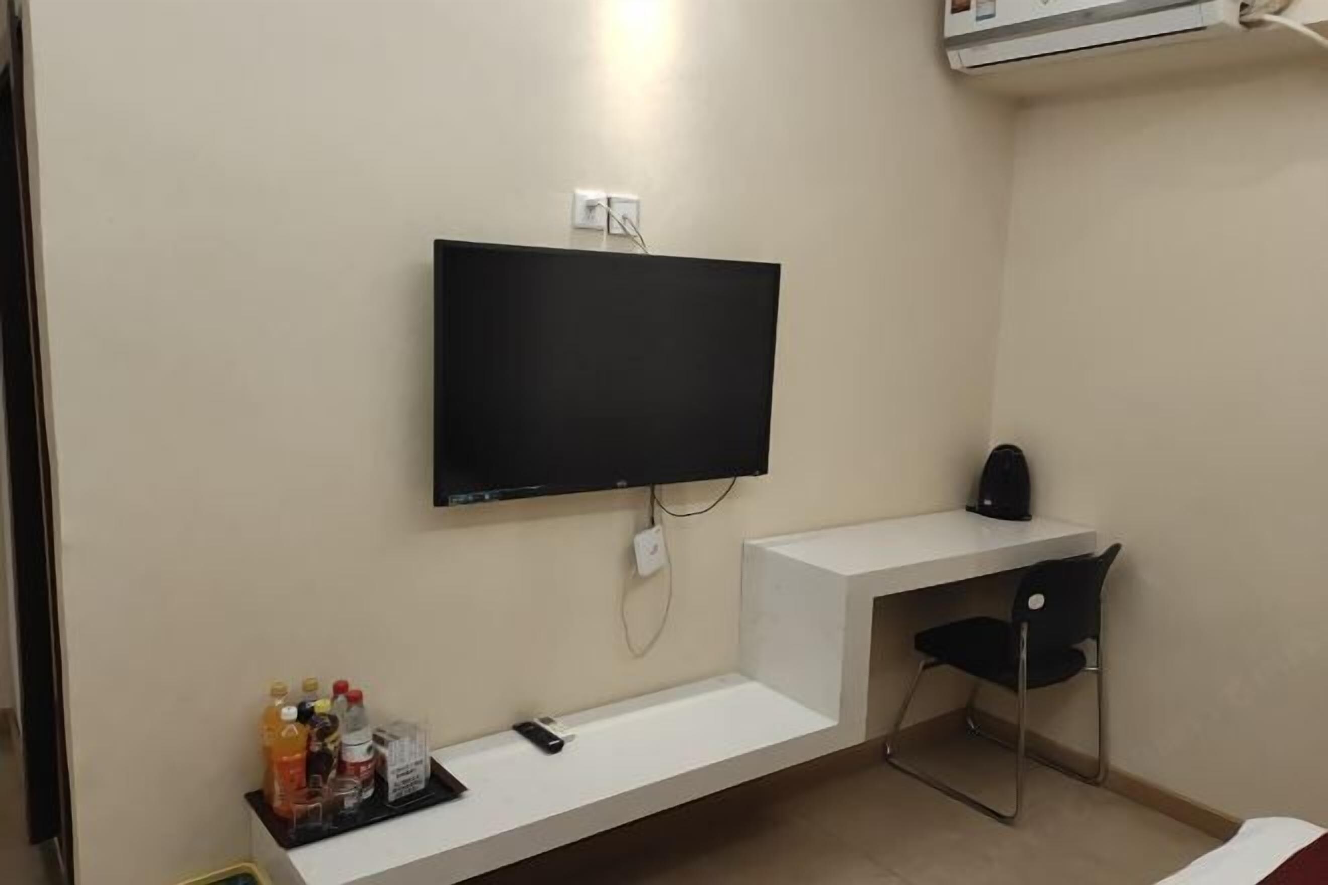 Ein Hotelzimmer mit Wand-Fernseher, einem weißen Schreibtisch mit Stuhl und einer kleinen Ablage mit Flaschen und einer Fernbedienung.