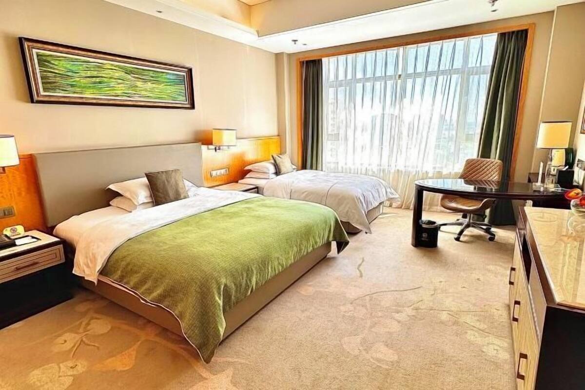 A hotel room with two beds, a desk, a chair, a lamp, and a window with curtains.