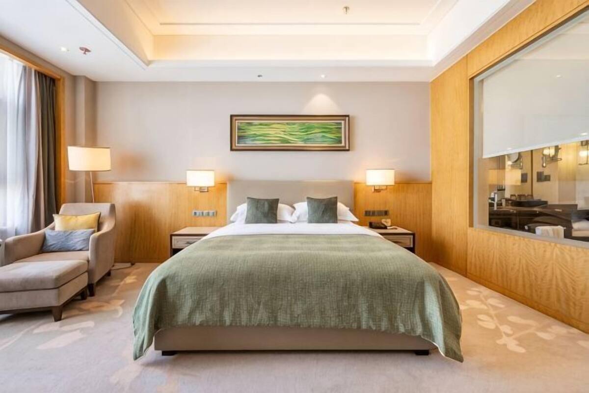 A hotel room with a bed, two bedside lamps, a chair, a large mirror, and a painting on the wall.