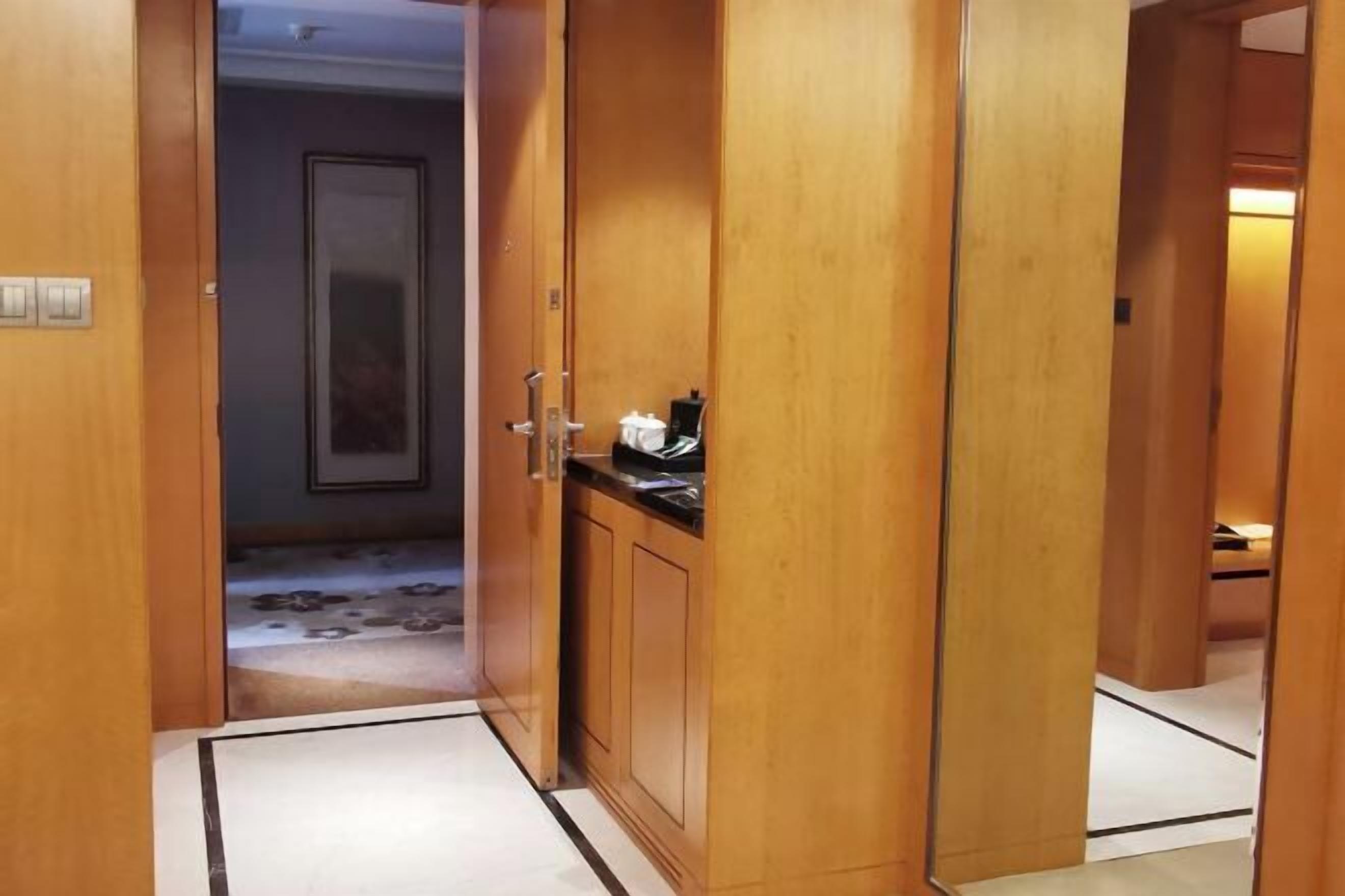 A wooden hallway with a door leading to a room. There is a small cabinet with a black countertop and a white object on it. The floor has a patterned design.
