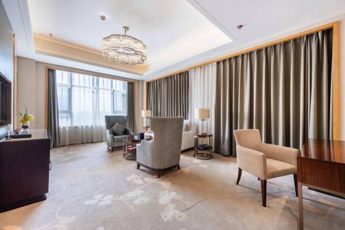 A hotel room with a large window, a chandelier, a sofa, a chair, and a desk.