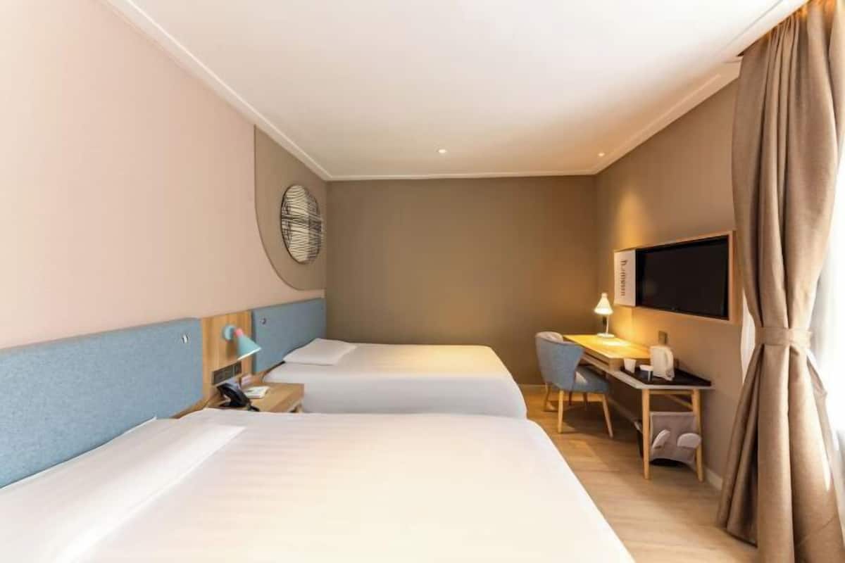 A hotel room with two beds, a desk, a chair, a TV, and a wall-mounted lamp.
