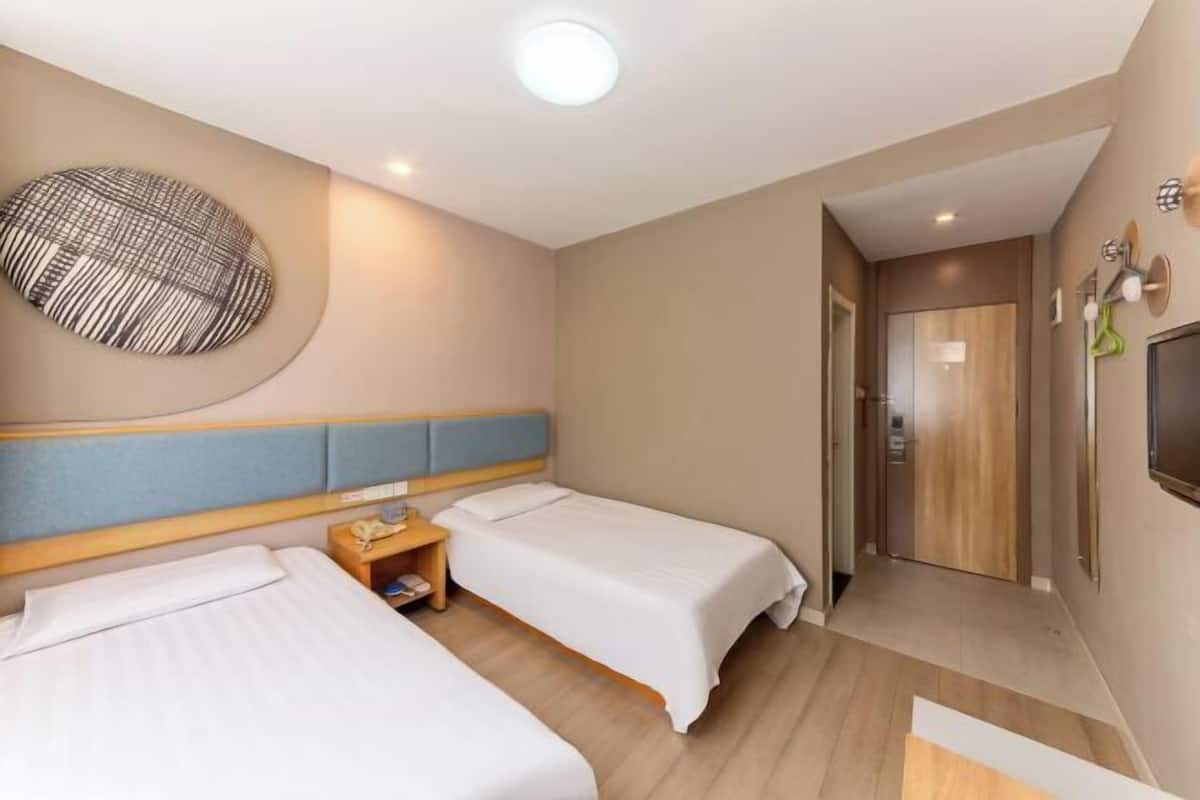 A hotel room with two beds, a round wall decoration, a wooden headboard, a bedside table with a lamp, and a television mounted on the wall.