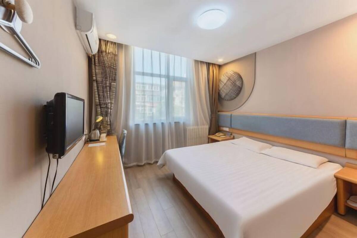 A hotel room with a bed, a desk with a television, a wall-mounted fan, and a window with curtains.