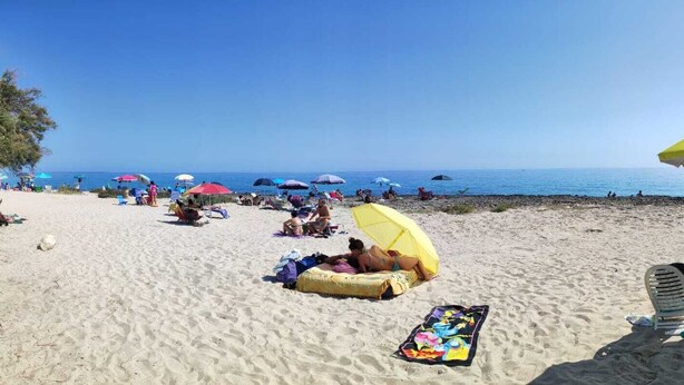 Ein Strand mit badenden Personen unter Sonnenschirmen, einem Liegestuhl und einer Kühlbox im Sand.