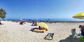 Una playa con gente tomando el sol bajo sombrillas, una silla de playa y una hielera en la arena.