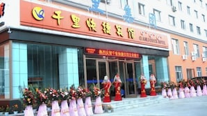A hotel entrance with a red sign, large windows, and a row of flower arrangements outside. - Qianli Express Hotel (Jiamusi)
