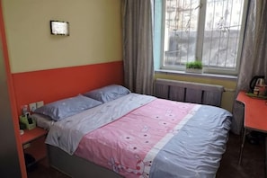 A single bed with a pink and blue bedspread, a nightstand with a lamp, and a window with curtains. - Qianli Express Hotel (Jiamusi)