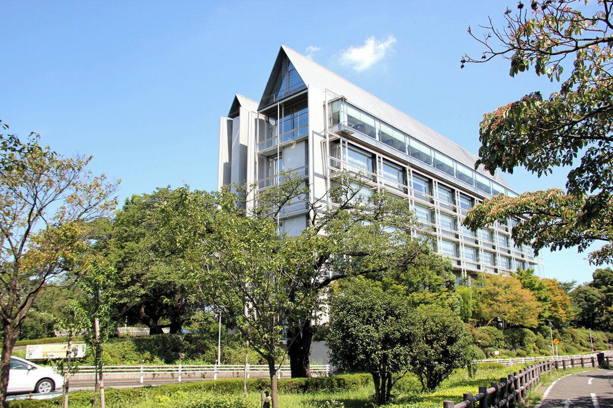 A modern building with a triangular roof and large windows, surrounded by lush greenery and a curved road.
