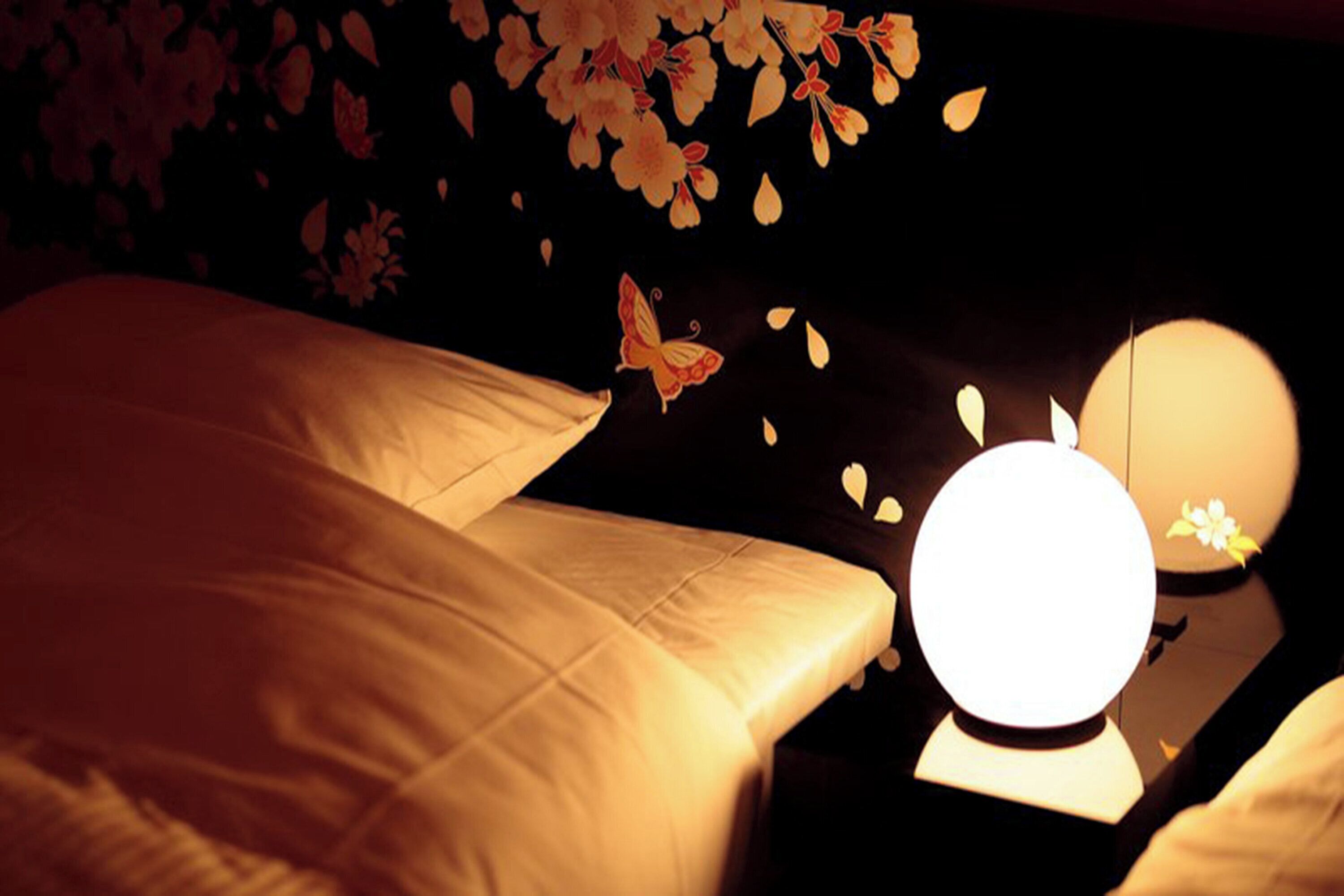Una habitación tenuemente iluminada con una cama, una mesita de noche con una lámpara y una pared decorada con motivos florales.