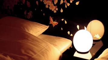 Una habitación tenuemente iluminada con una cama, una mesita de noche con una lámpara y una pared decorada con motivos florales.