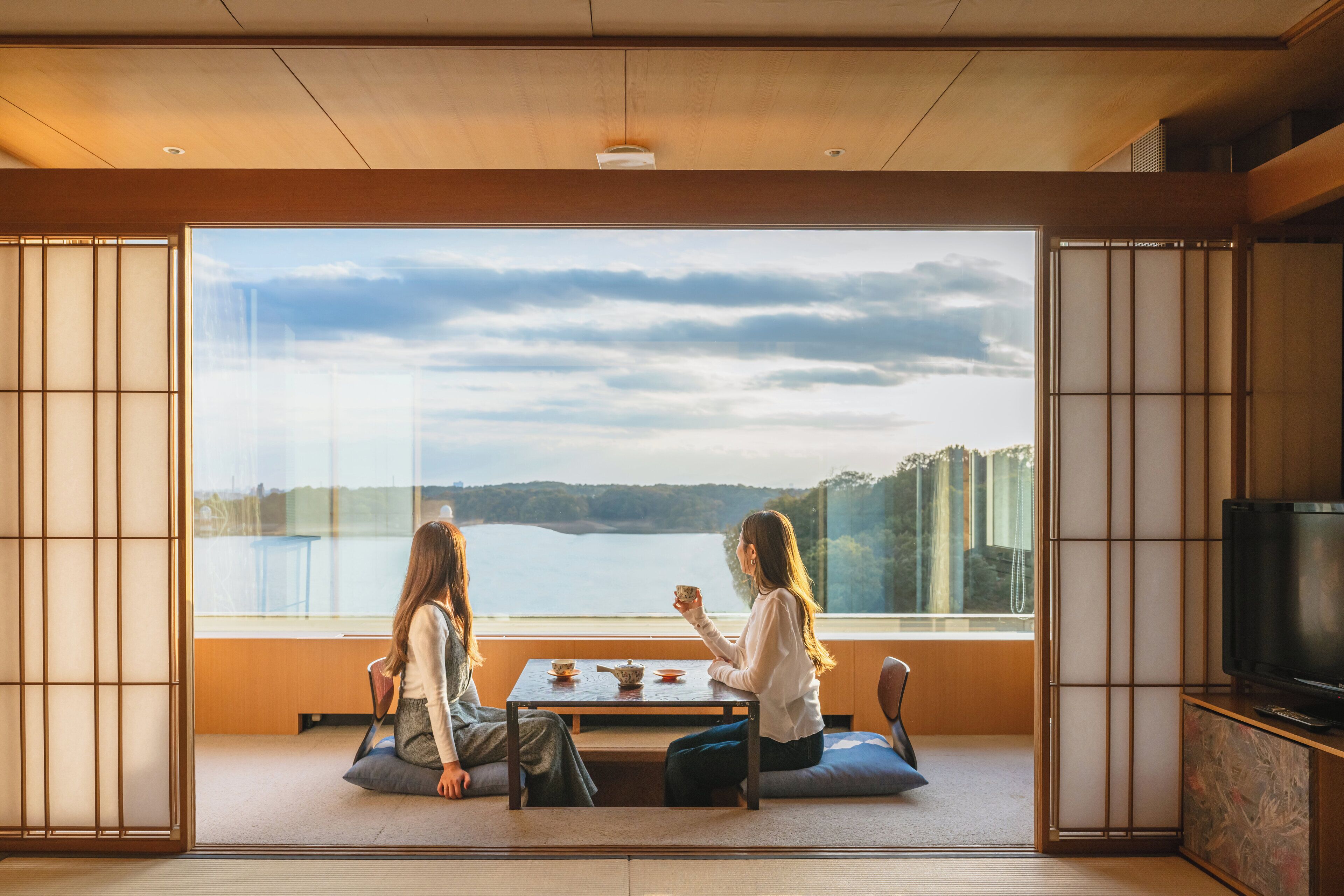 Une pièce au sol recouvert de tatamis, avec une table basse et une grande fenêtre donnant sur un lac et des bâtiments situés au loin.