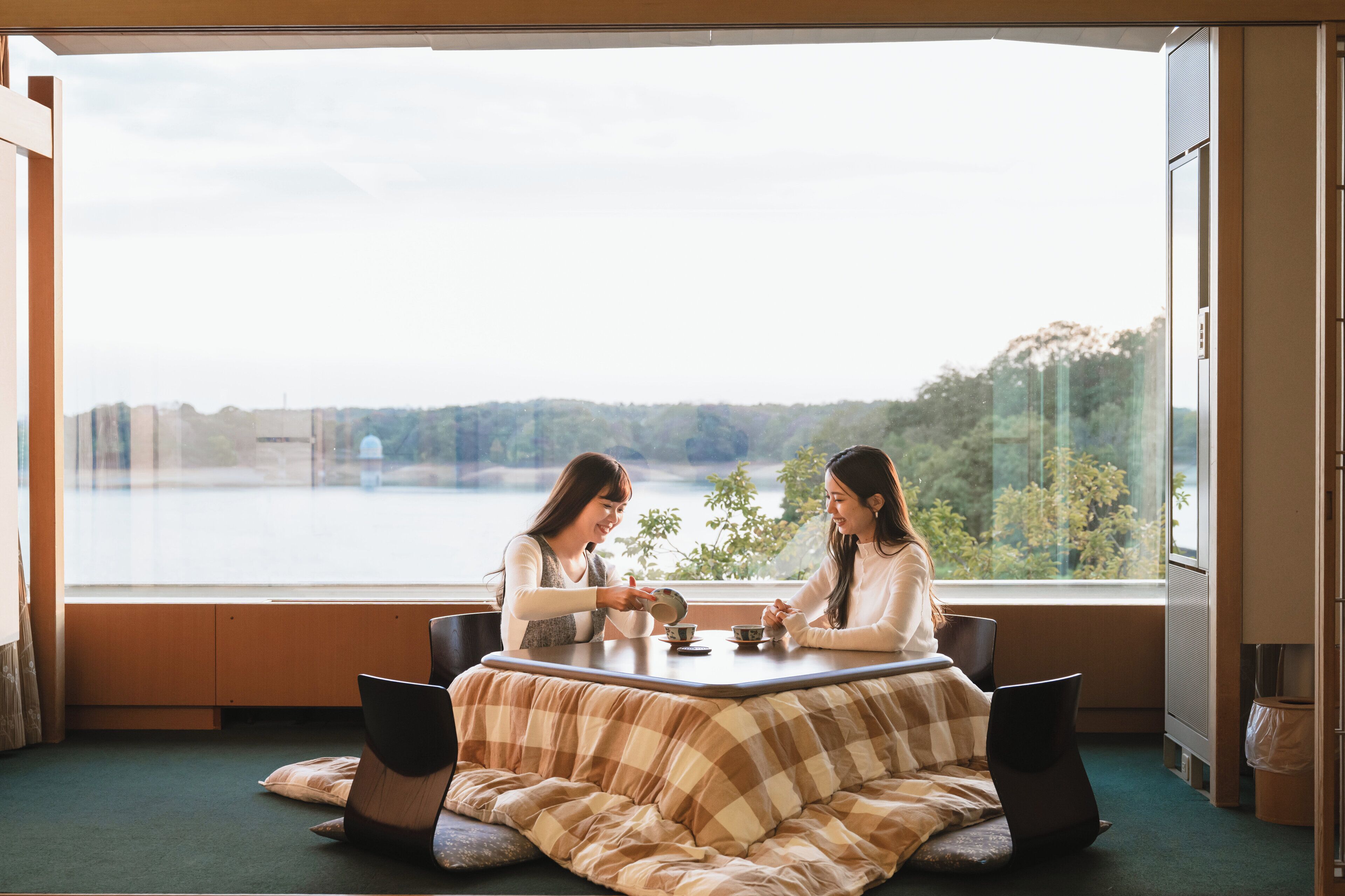 A room with a large window overlooking a lake, a cozy seating area with a checkered blanket, and a table with cups and saucers.