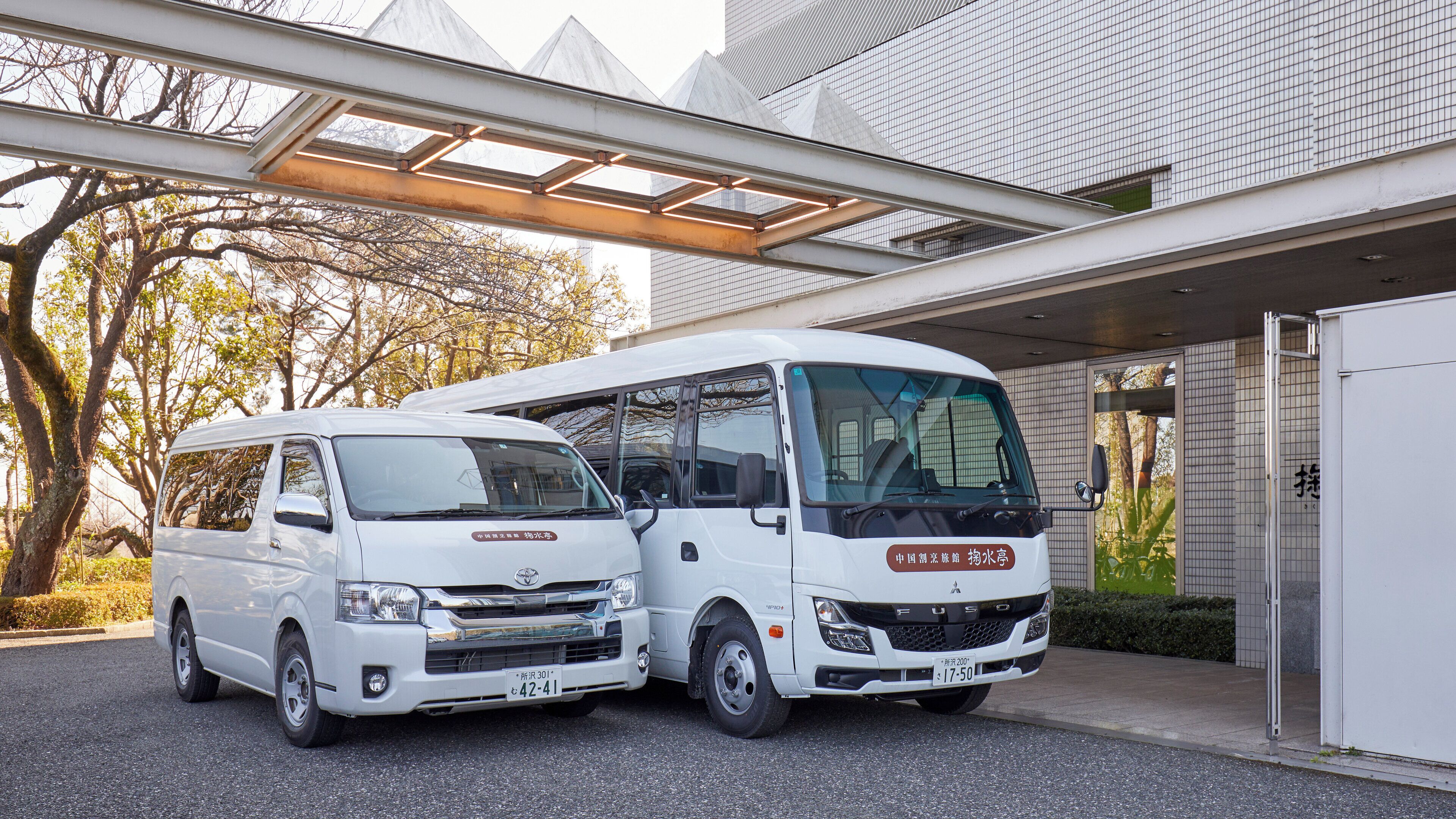 A white van and a white bus parked in front of a building with a modern design, including a glass canopy structure.