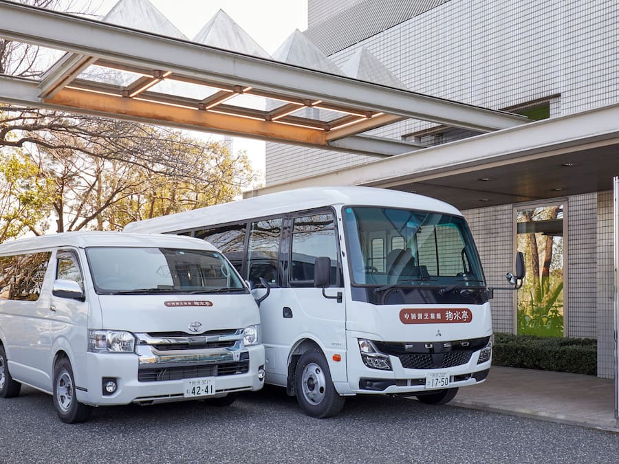 A white van and a white bus parked in front of a building with a modern design, including a glass canopy structure.