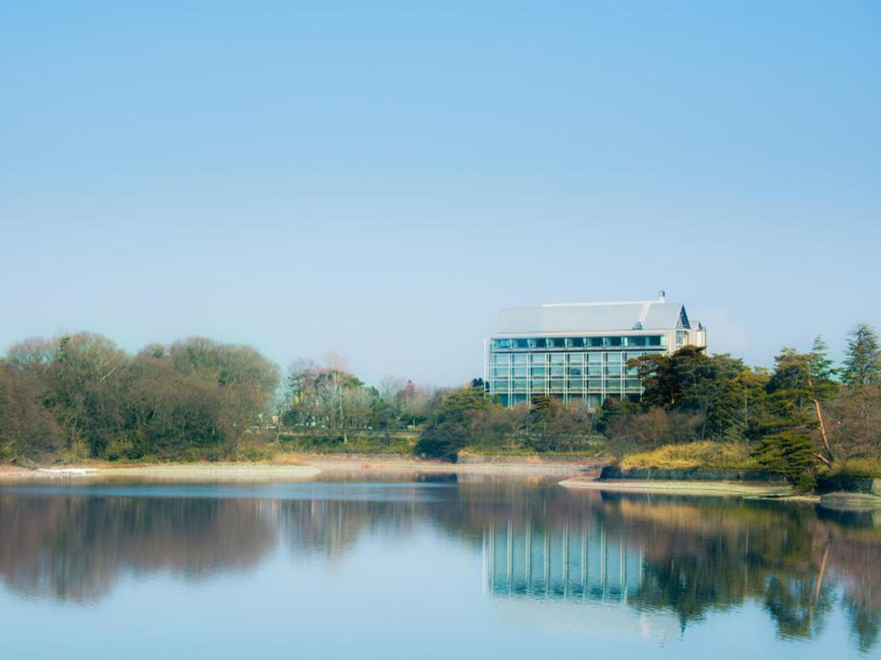 A modern building with a glass facade, situated by a calm lake with trees in the background.