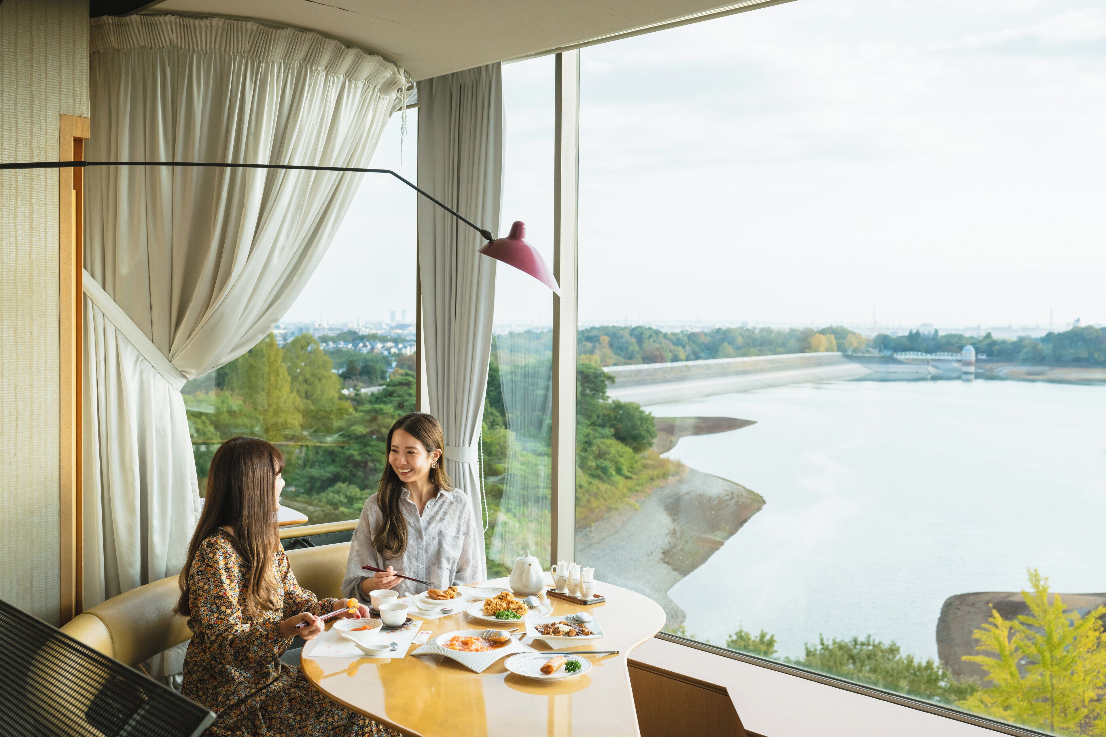 A dining area with a table set for two, a view of a river, and a cityscape in the background.