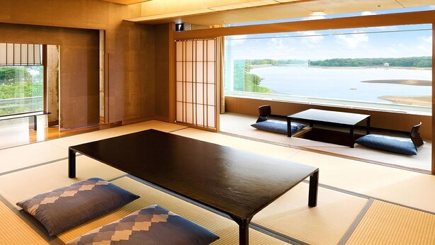 A modern room with tatami flooring, a low table, and cushions. A large window offers a view of a river and greenery. - Kikusuitei (Higashiyamato)