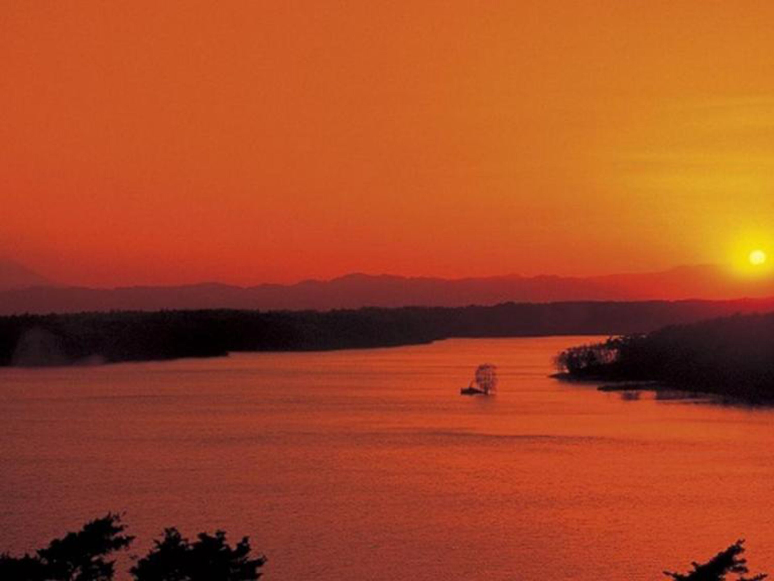 Un atardecer sobre un lago tranquilo con un barco a lo lejos y montañas al fondo.