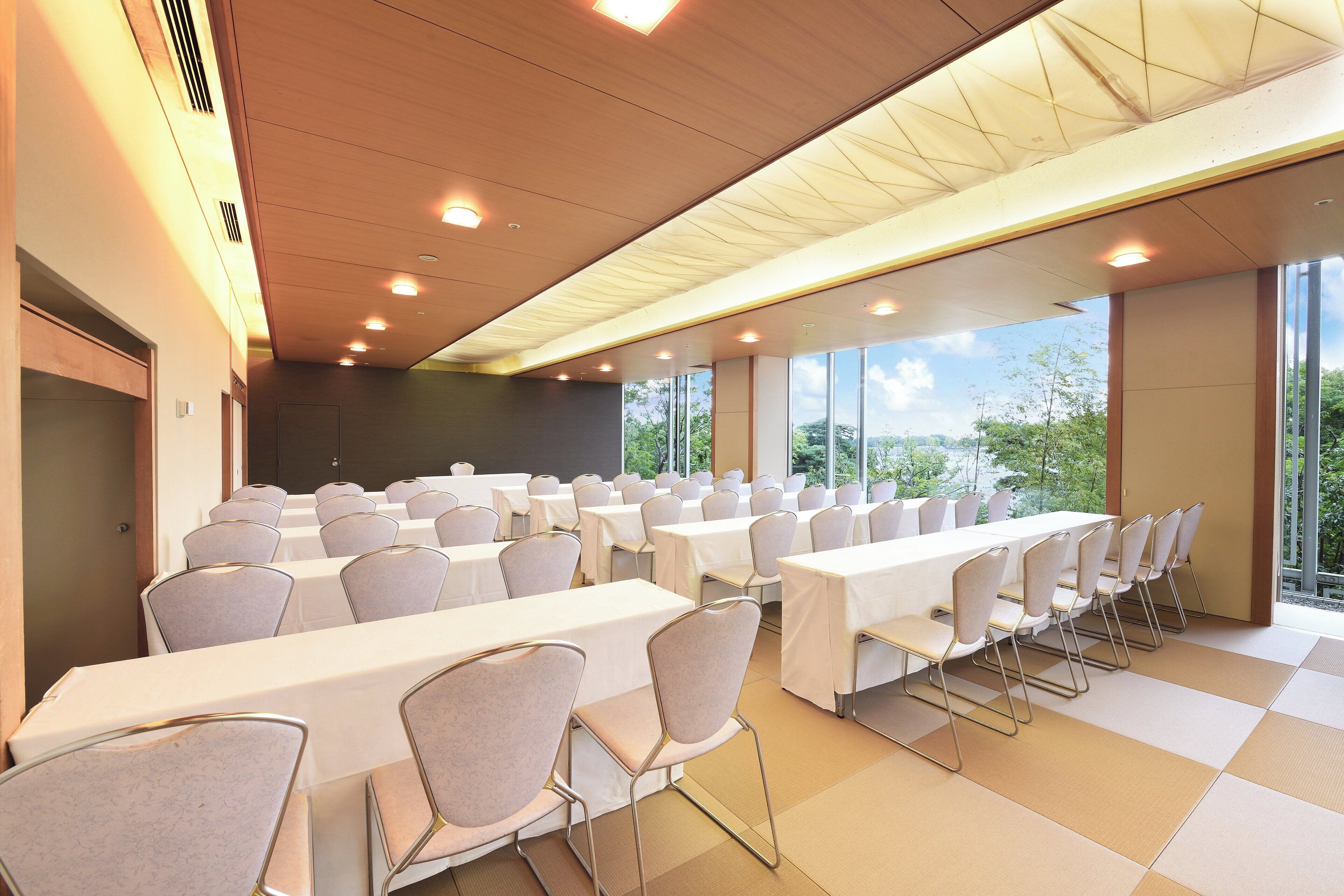 A conference room with multiple tables and chairs, large windows, and a high ceiling with recessed lighting.