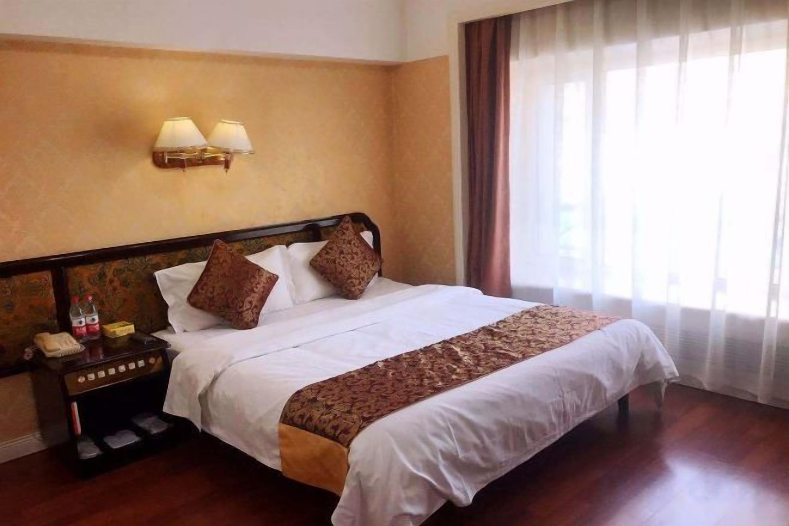 A hotel room with a large bed, a nightstand with a lamp, and a view of the outdoors through a window.