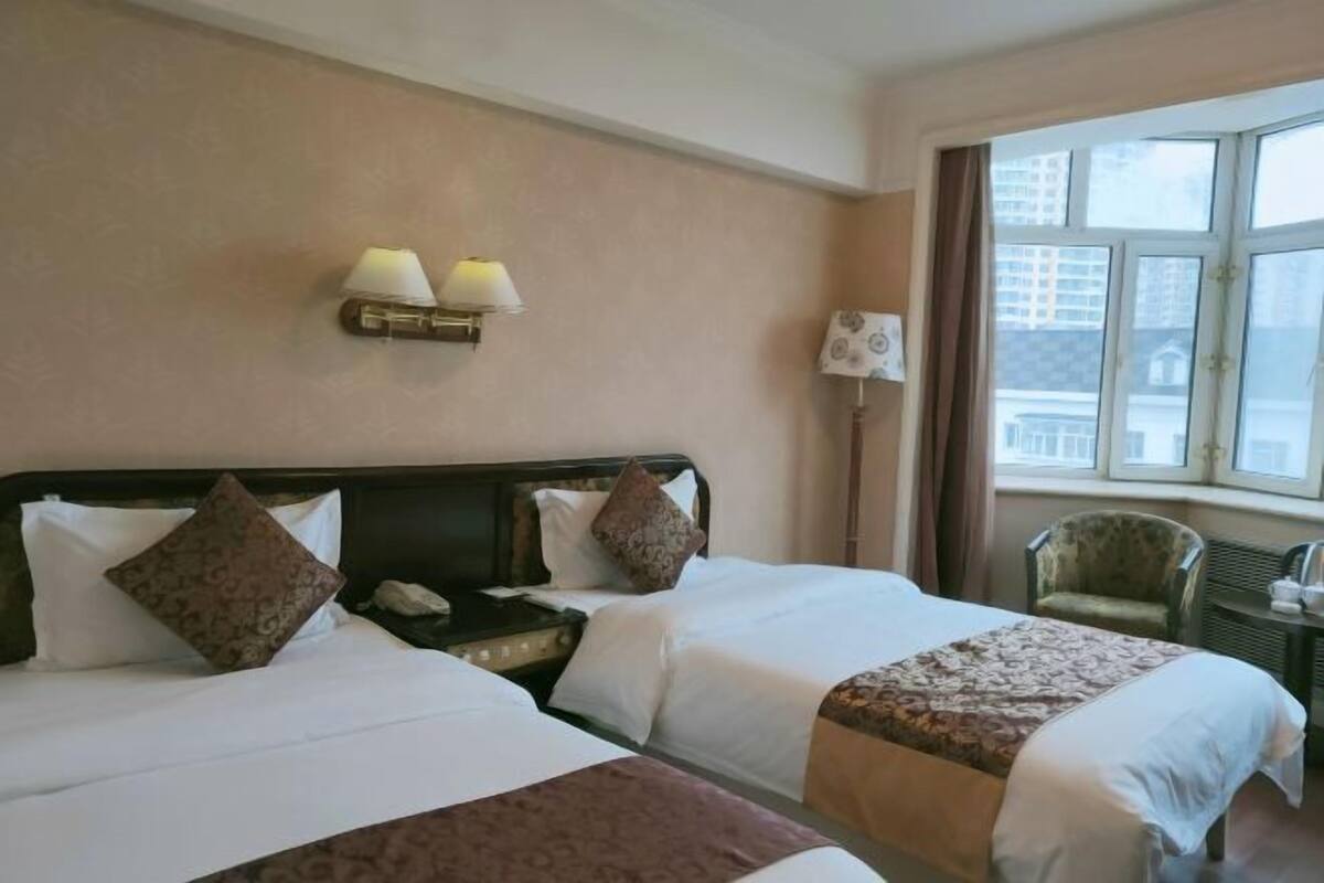 Une chambre d’hôtel avec deux lits, une table de chevet, une lampe, une chaise et une fenêtre donnant sur des immeubles.