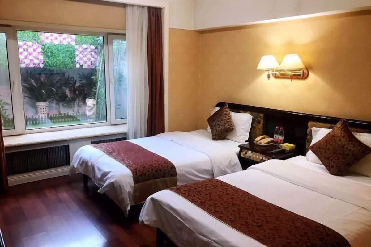 Une chambre d’hôtel avec deux lits, une fenêtre donnant sur un paysage verdoyant, une lampe fixée au mur et une table de chevet avec un téléphone et une bouteille.