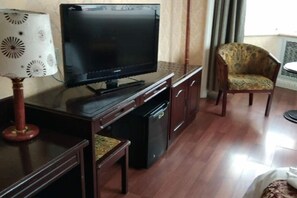 A room with a flat-screen TV on a wooden stand, a small table with a lamp, a patterned armchair, and a cabinet. - Fushun Tiantian Hotel (Mudanjiang High-speed Railway Wanda Branch) (Mudanjiang)