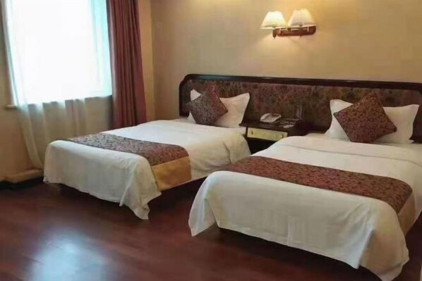 Dos camas individuales con sábanas blancas y cabeceras marrones, una mesita de noche con lámpara, y una ventana con cortinas.