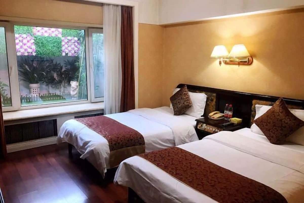 Une chambre d’hôtel avec deux lits, une fenêtre donnant sur un espace verdoyant, une lampe fixée au mur et une table de chevet avec un téléphone et une bouteille.