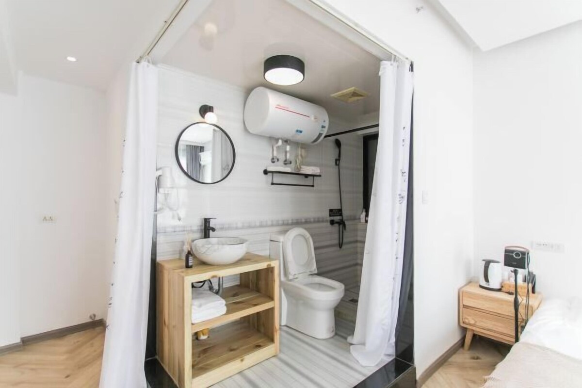 A compact bathroom with a toilet, a sink with a mirror, a shower, and a bedside table with a coffee maker.
