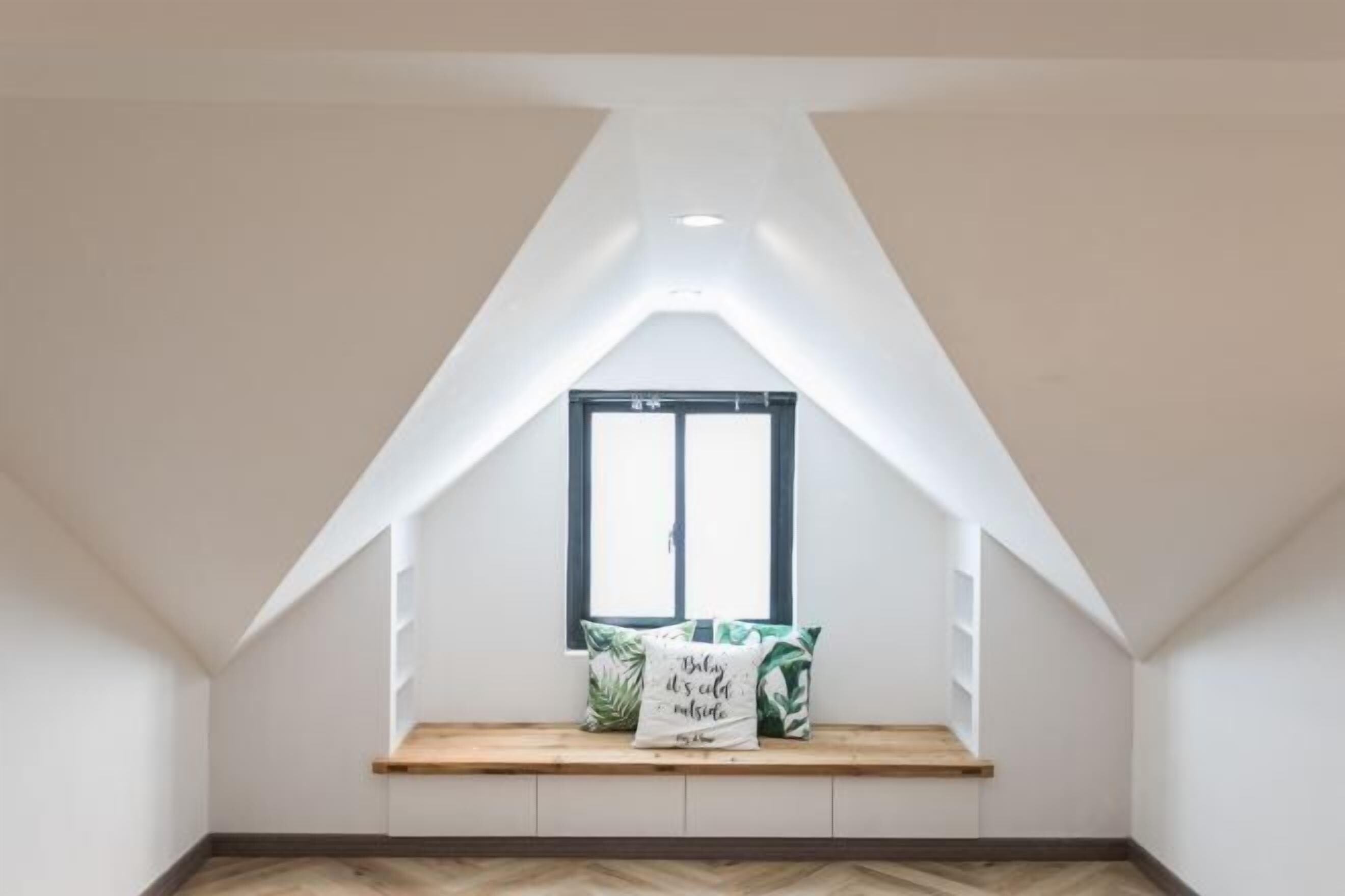 A room with a sloped ceiling, a window with a black frame, a wooden bench with cushions, and a herringbone-patterned floor.
