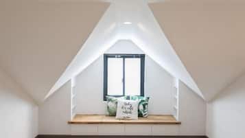 A room with a sloped ceiling, a window with a black frame, a wooden bench with cushions, and a herringbone-patterned floor.