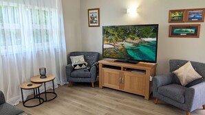 A living room with a large flat-screen TV, two grey armchairs, a wooden TV stand, a small round table with a candle, and sheer white curtains. - Jack's Beach Shack by Experience Jervis Bay (Huskisson)