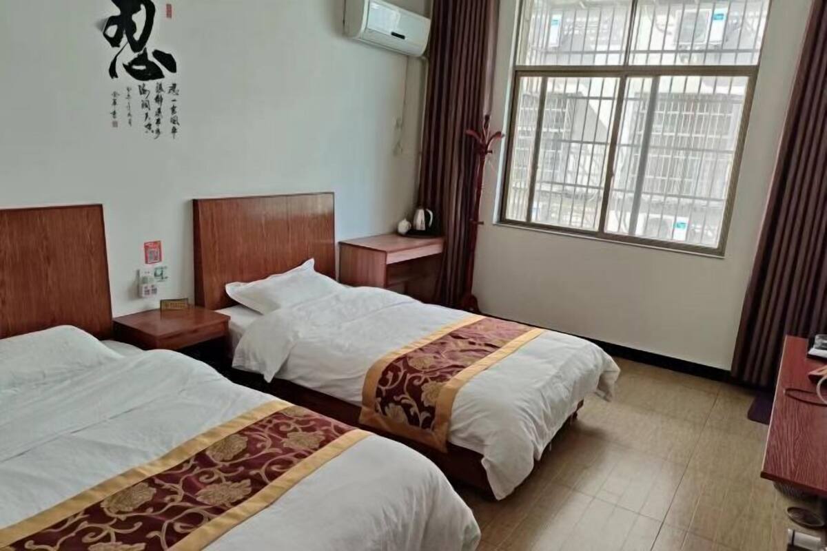A hotel room with two beds, a wooden headboard, a nightstand, a window with curtains, and a wall-mounted air conditioner.