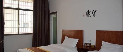A hotel room with two beds, a window with curtains, a wooden headboard, and a small bedside table with a radio.