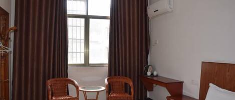 Habitación de hotel con cama, dos sillas de mimbre, una mesa redonda pequeña, un escritorio con silla y una ventana con cortinas.