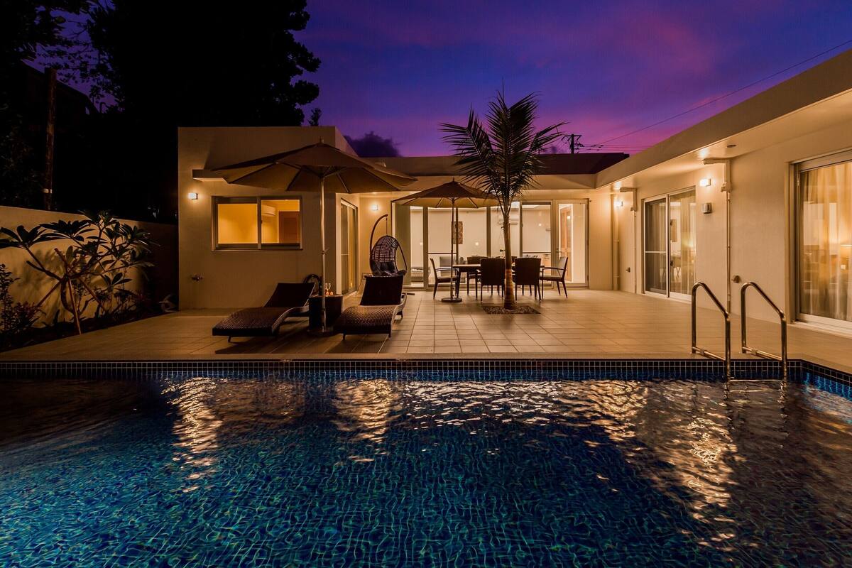 A modern outdoor pool area with lounge chairs and a dining table, surrounded by a covered patio and a glass-enclosed room.