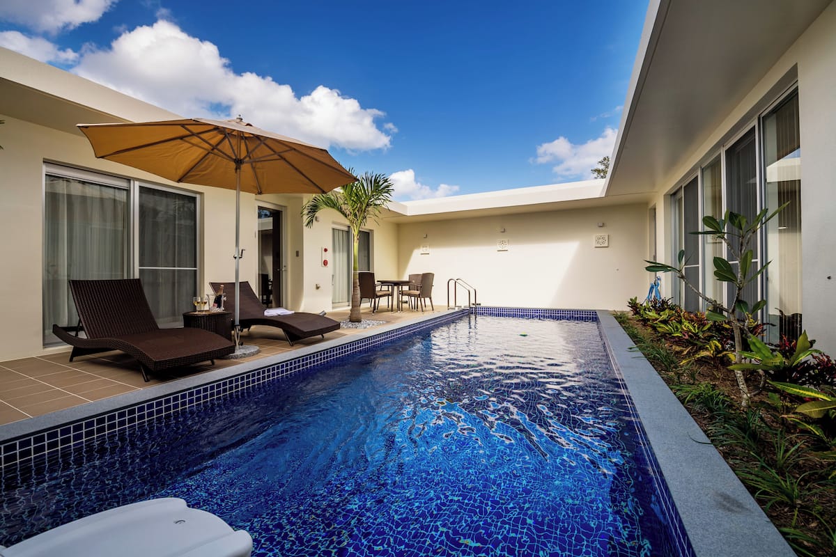 A swimming pool with lounge chairs and an umbrella, surrounded by a patio area with plants and a view of the sky.