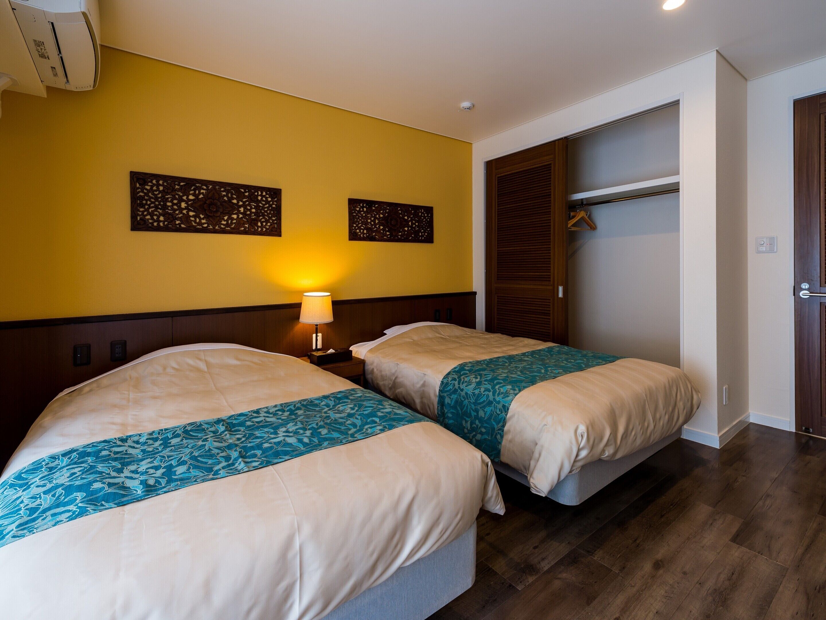 A hotel room with two beds, a nightstand with a lamp, and a wardrobe.