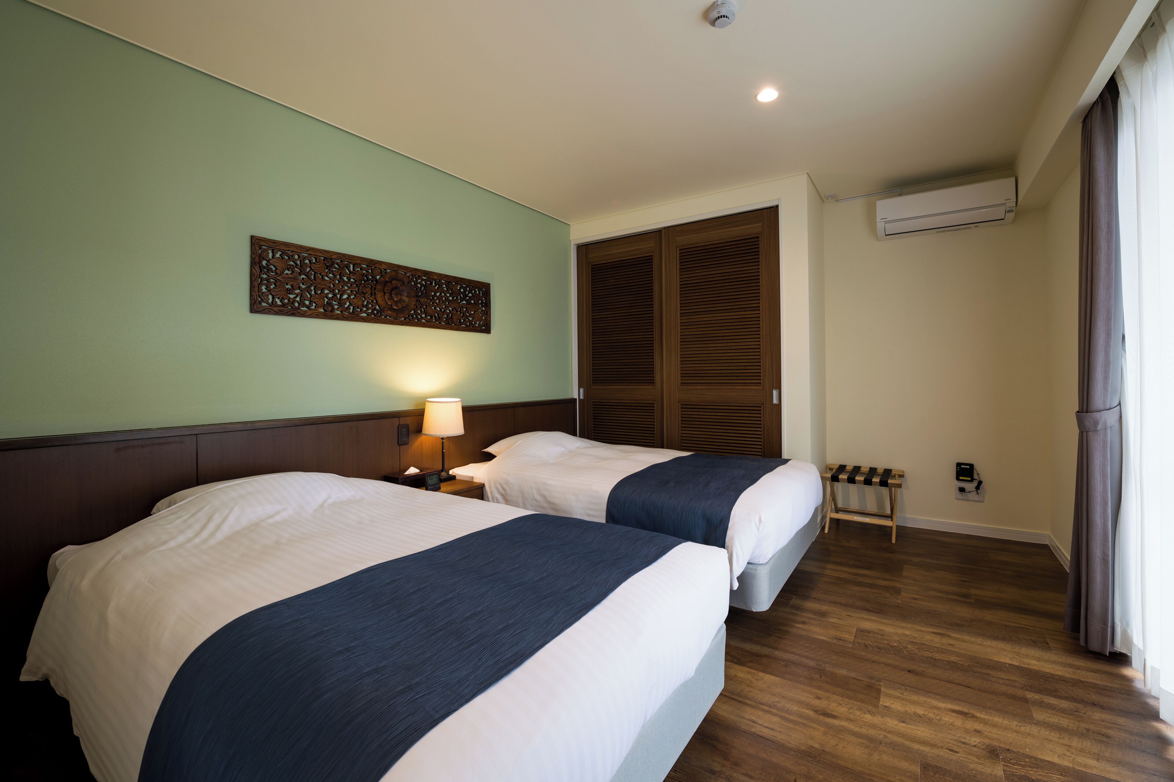 A hotel room with two beds, a nightstand with a lamp, a decorative wall piece, a wooden bench, and a window with curtains.