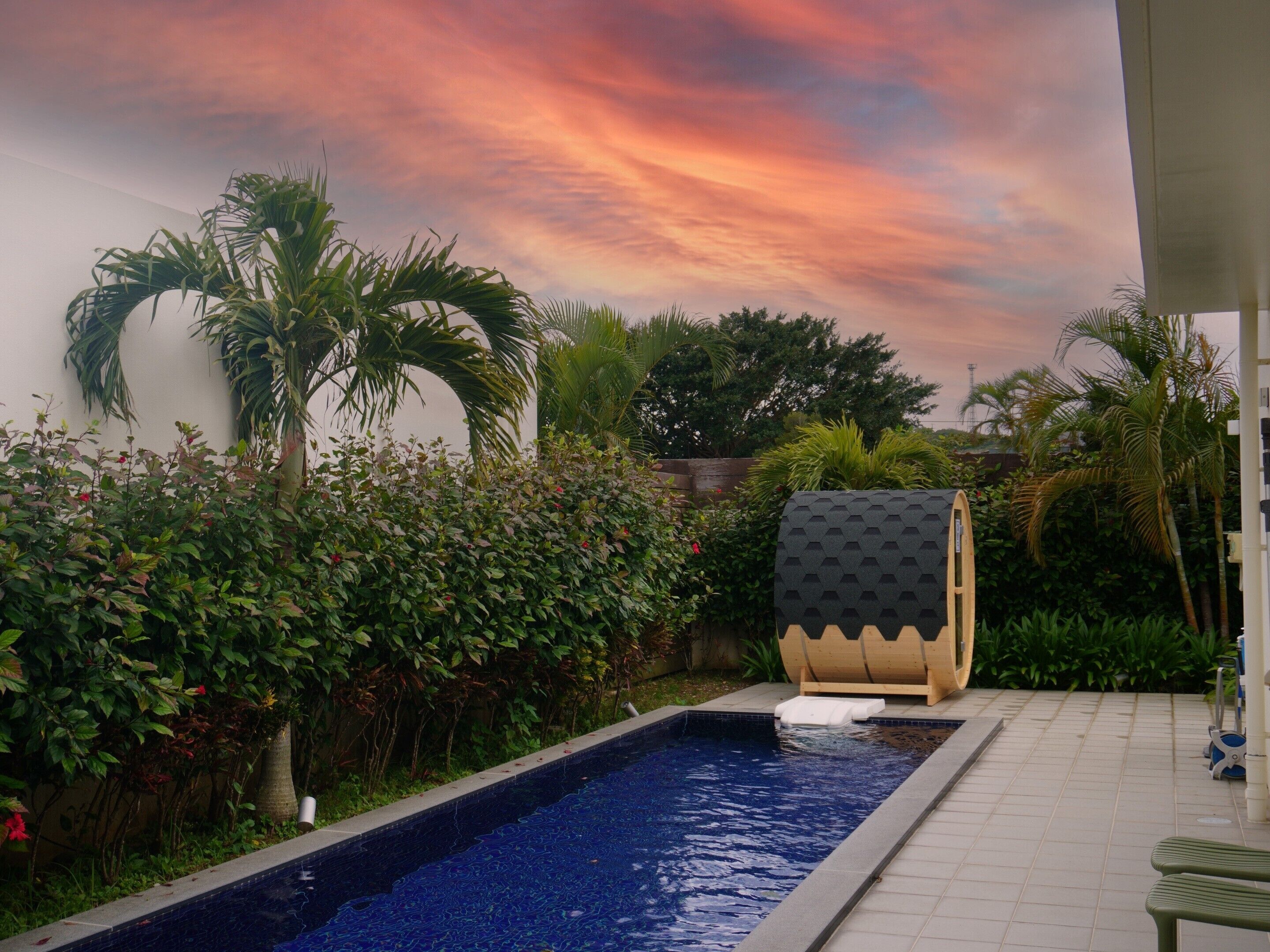 A swimming pool with a tiled edge, surrounded by lush greenery and a wooden structure with a checkered pattern.