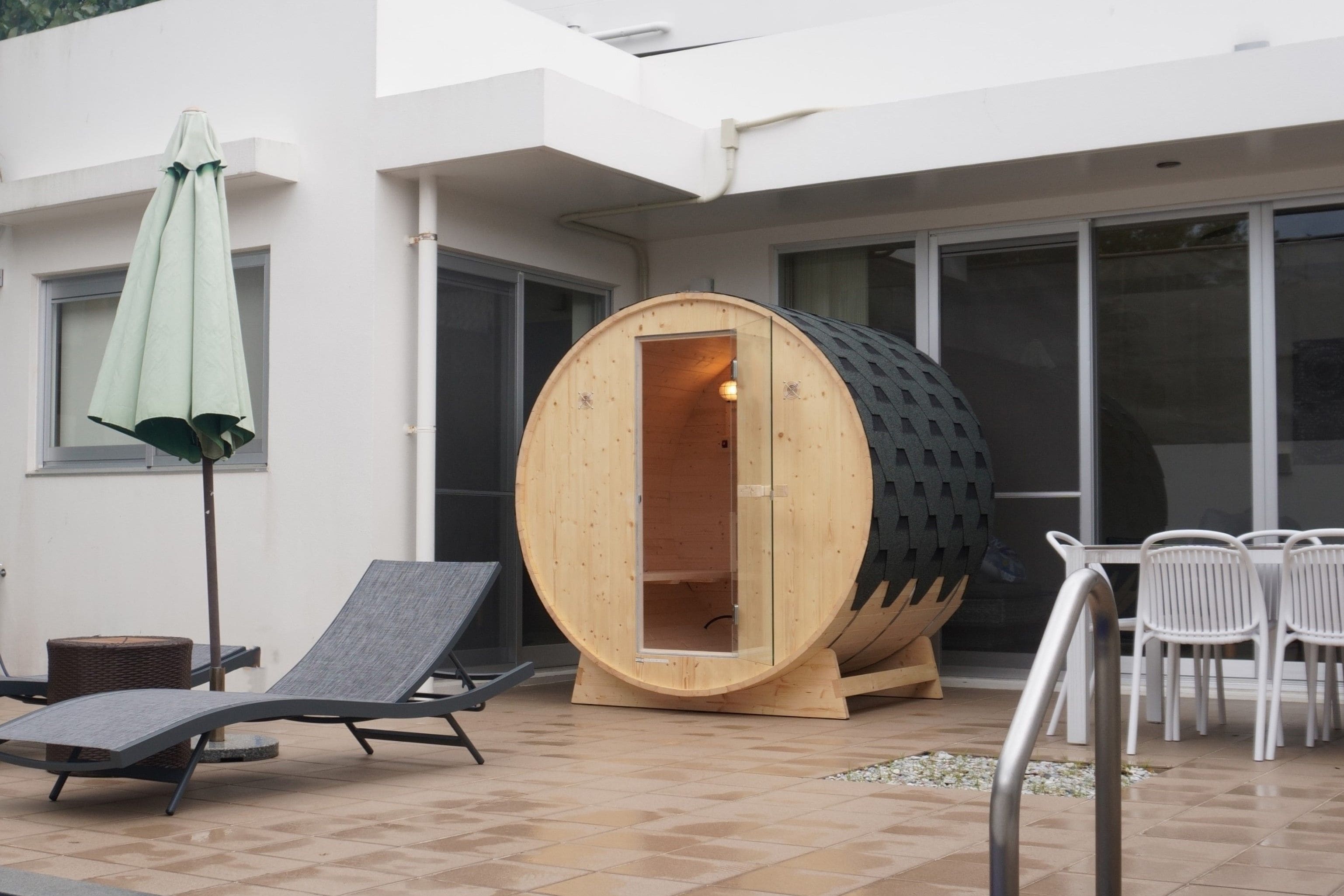 A sauna with a wooden exterior and a glass door, a lounge chair, a small table, and a white umbrella.